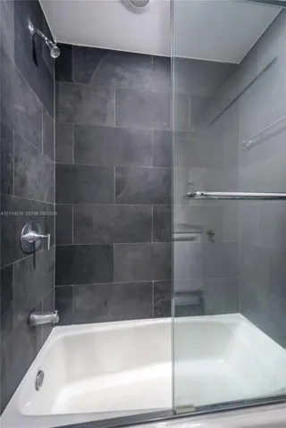 Bathtub with a glass enclosure, featuring large-format dark stone wall tile, chrome shower head, and wall-mounted chrome hardware