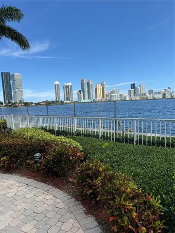 Waterfront property featuring a paved stone walkway, landscaped shrubbery, a white perimeter fence, and a skyline backdrop