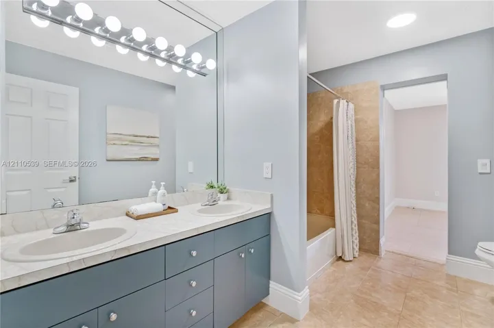 Dual-sink vanity with a solid-surface countertop and recessed basins, expansive wall-mounted vanity mirror, multi-bulb overhead lighting, and tub-shower combination featuring neutral-toned tile surround