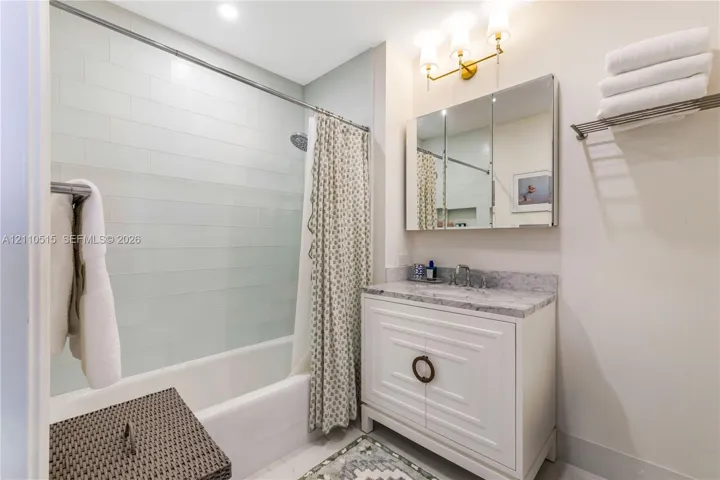 Bathroom featuring a vanity with stone-finish countertop, tri-fold medicine cabinet, three-light fixture, tub-shower combination with tile surround, and built-in wall shelf
