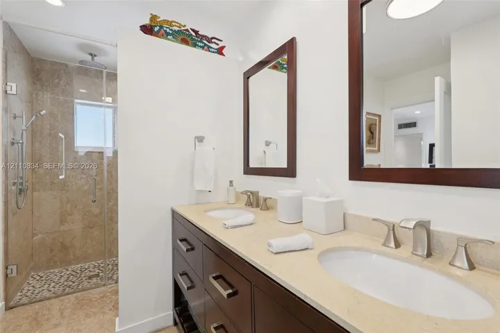 Double vanity with stone countertop, dark wood-finish cabinetry, frameless glass shower enclosure, stone-tiled shower wall, and integrated shower rainfall fixture