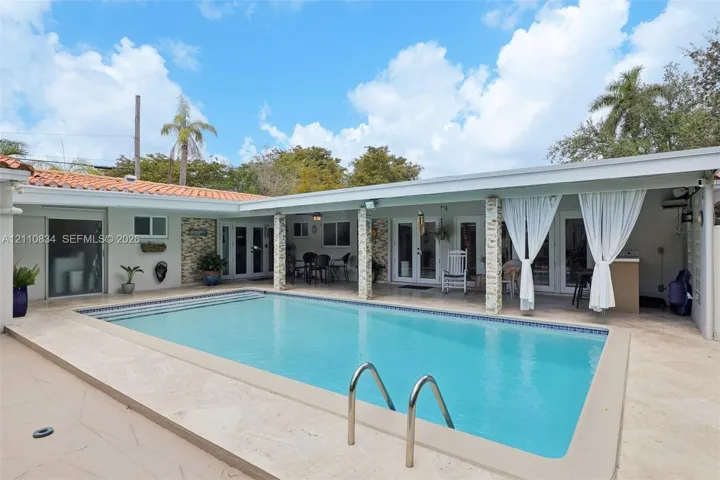 In-ground swimming pool with stone-tiled decking and covered patio area featuring stone-clad support columns and light-toned exterior finish
