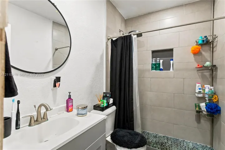 Bathroom featuring a vanity with a white countertop and integrated sink, matte-finish faucet, circular wall mirror, and a shower area with tiled walls and a recessed niche