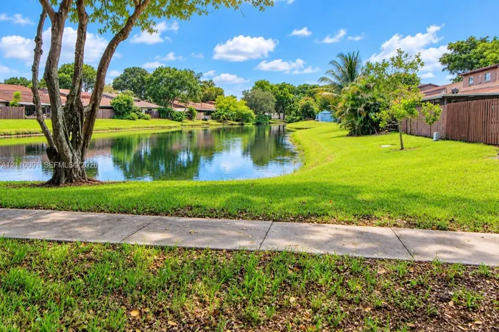 Waterfront grounds featuring a concrete walking path, mature trees, manicured lawn, and perimeter wood fencing