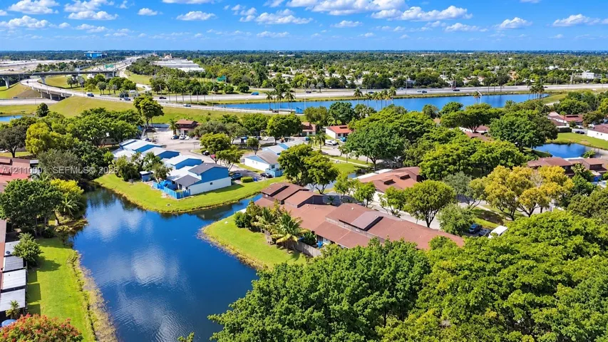 Aerial perspective of a multi-unit residential property featuring waterfront positioning, expansive green space, mature landscaping, and brown shingle roofing