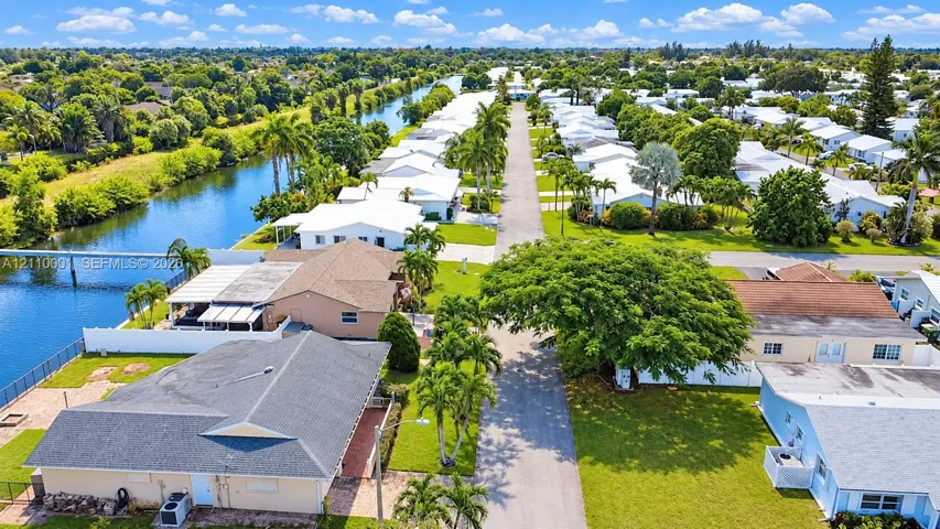 Aerial perspective of a residential street featuring waterfront access, paved thoroughfare, manicured landscaping, mature canopy trees, and established perimeter fencing