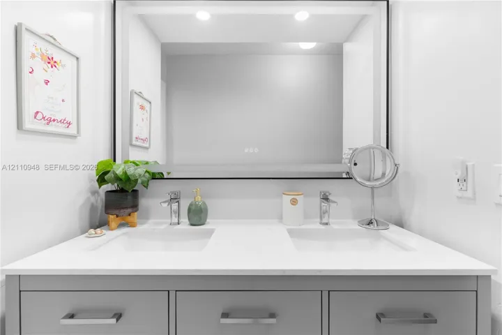 Double-sink vanity featuring a quartz countertop, soft-grey shaker-style cabinetry, polished chrome faucets, and a large integrated LED-backlit mirror with a black frame