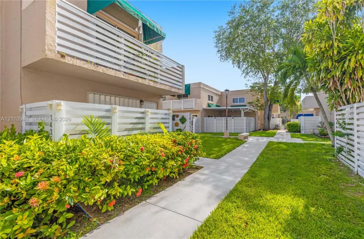 Exterior featuring a concrete walkway, white slatted privacy fencing, second-story balcony with fabric awning, and mature tropical landscaping
