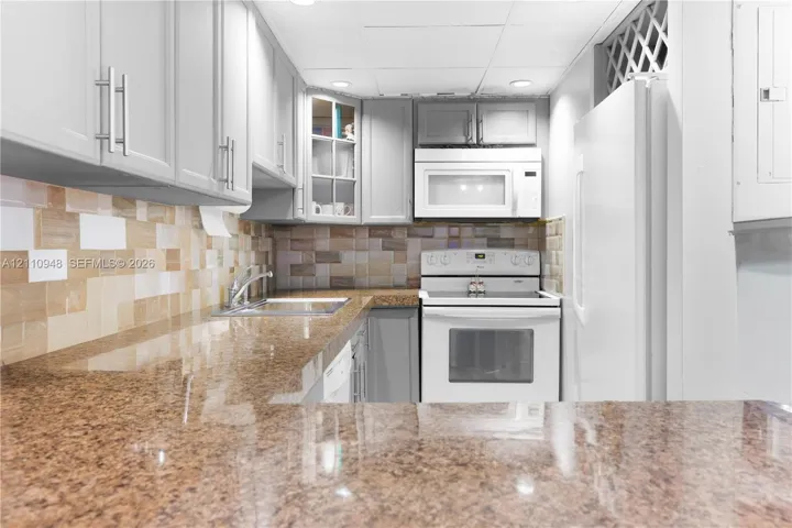 Kitchen featuring stone countertops, stone tile backsplash, light grey cabinetry with glass-front accents, and white appliances