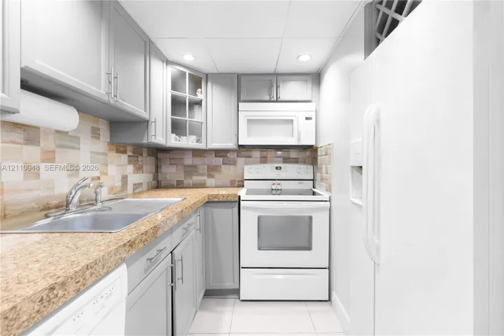 Kitchen featuring shaker-style cabinetry, multi-tonal tile backsplash, stone-surface countertops, dual-basin stainless steel sink, and white appliances