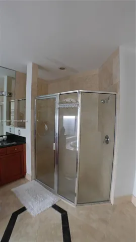 Corner shower enclosure with a metallic frame, neutral stone-finish wall tile, vanity with dark stone countertop, integrated mirror, and patterned tile flooring