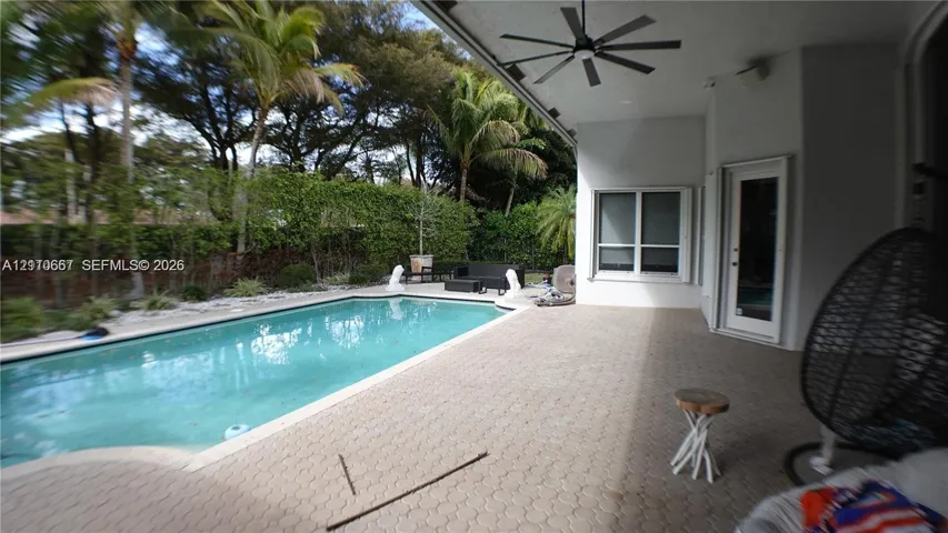 Covered patio with a ceiling fan, concrete paver decking, and an in-ground swimming pool enclosed by mature perimeter landscaping