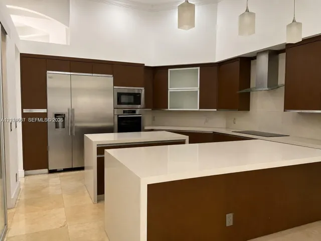 Kitchen featuring flat-panel dark wood cabinetry, waterfall-edge stone countertops, stainless steel appliances, glass-front upper cabinets, and pendant lighting