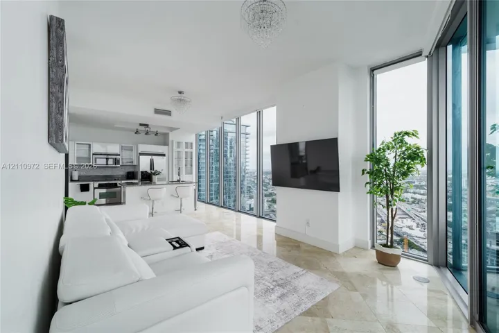 Floor-to-ceiling windows, polished stone tile flooring, stainless steel appliances, white cabinetry, and crystal-style chandelier lighting