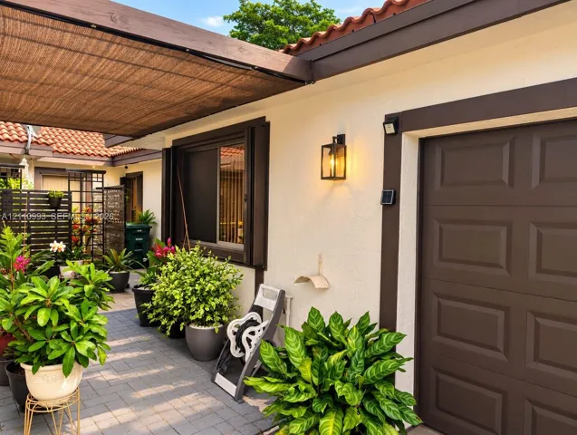 Exterior courtyard featuring paver flooring, a tensioned shade canopy, stucco wall finish, dark-toned trim, and wall-mounted lantern-style lighting