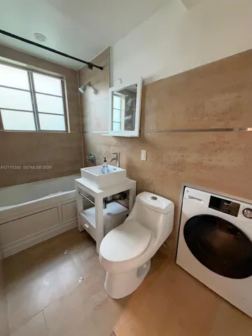 Bathroom featuring large-format wall and floor tile, a vessel sink on a white open-frame vanity, a built-in soaking tub, and an integrated laundry appliance