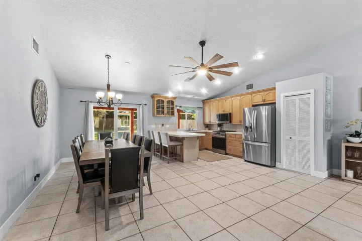 Open-concept kitchen and dining area featuring vaulted ceilings, ceramic tile flooring, natural wood cabinetry, stainless steel appliances, and a central island with bar seating