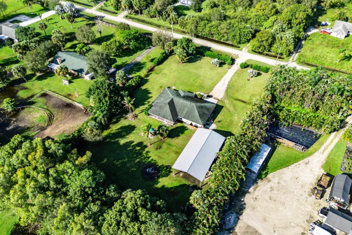 Aerial perspective of a multi-structure property featuring a residence with a dark-tone shingled roof, a detached metal-roofed structure, extensive grassy acreage, mature perimeter trees, and a gravel driveway