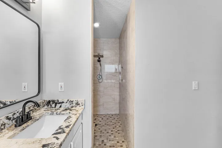 Vanity with stone countertop and undermount sink, matte black faucet, wall-mounted mirror, and walk-in shower with stone tile wall surfacing and mosaic tile flooring
