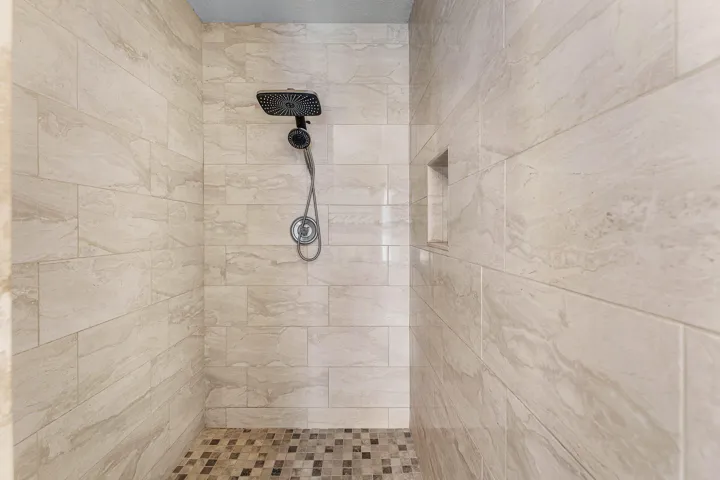 Walk-in shower featuring large-format stone-finish wall tile, a square rainfall showerhead, a hand-held shower sprayer, a built-in recessed wall niche, and mosaic tile flooring