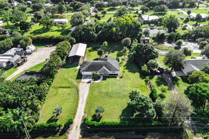 Aerial view of a property featuring a single-story residence, a detached metal-roofed structure, a gravel driveway, and expansive lawn areas with mature trees