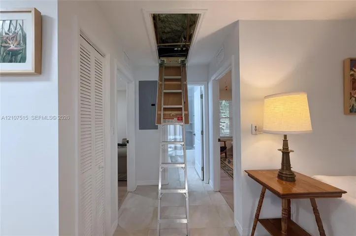 Hallway featuring attic access ladder, neutral tile flooring, and integrated electrical panel