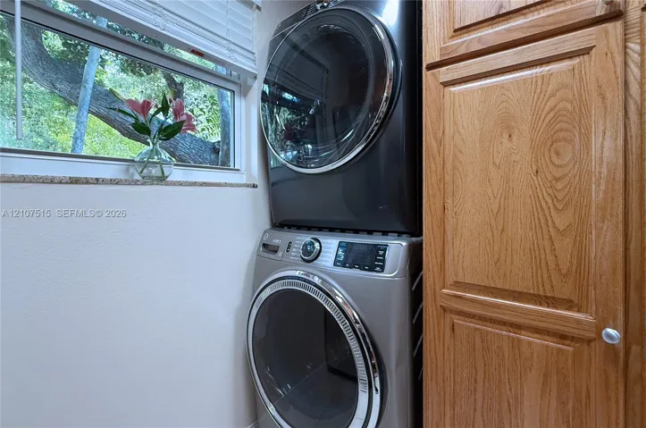 Stacked laundry center featuring a metallic finish washer and dryer, custom-built natural wood cabinetry, and a stone window sill