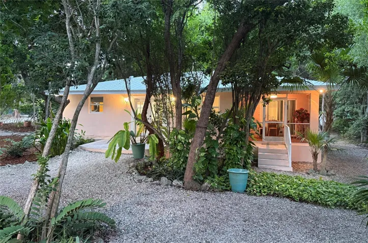 Single-story exterior featuring a metal roof, screened-in porch, concrete stairs, and a gravel ground surface surrounded by mature trees