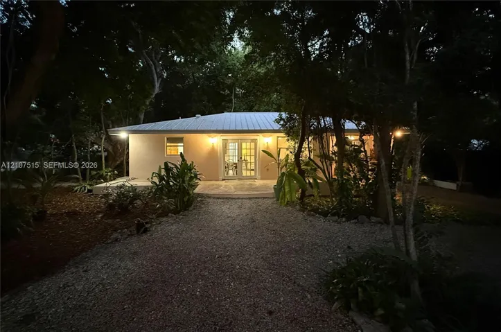 Single-story exterior featuring a metal roof, French doors, and a gravel driveway surrounded by mature trees