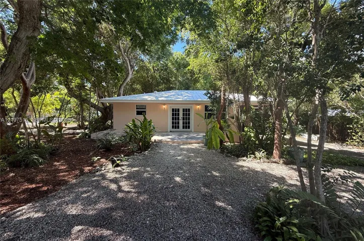 Single-story exterior featuring a metal roof, light-toned stucco facade, and double French doors, surrounded by mature trees and a gravel driveway