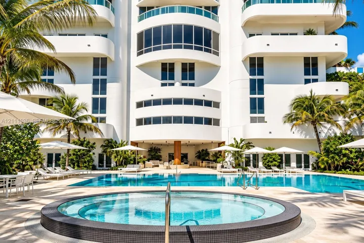 Multi-story exterior featuring curved balconies, floor-to-ceiling windows, a circular spa with tiled surround, a sprawling swimming pool, and stone paver decking