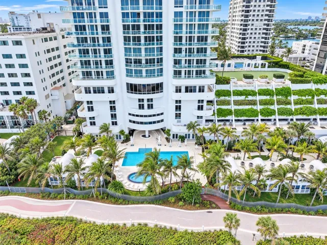 High-rise residential exterior featuring expansive glass balconies, a ground-level swimming pool, a separate spa, landscaped grounds with mature palm trees, and multi-level terraced gardens