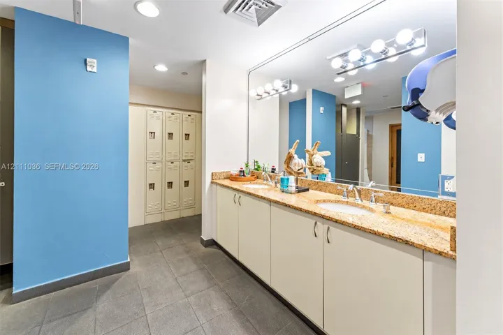 Dual-sink vanity with solid-surface countertop, floor-to-ceiling metal lockers, large-format tile flooring, and multi-bulb wall lighting