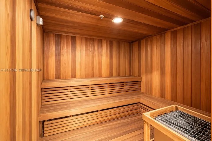 Sauna featuring tongue-and-groove wood paneling, multi-level built-in bench seating, recessed ceiling lighting, and a stone-filled heating unit with a wooden safety enclosure