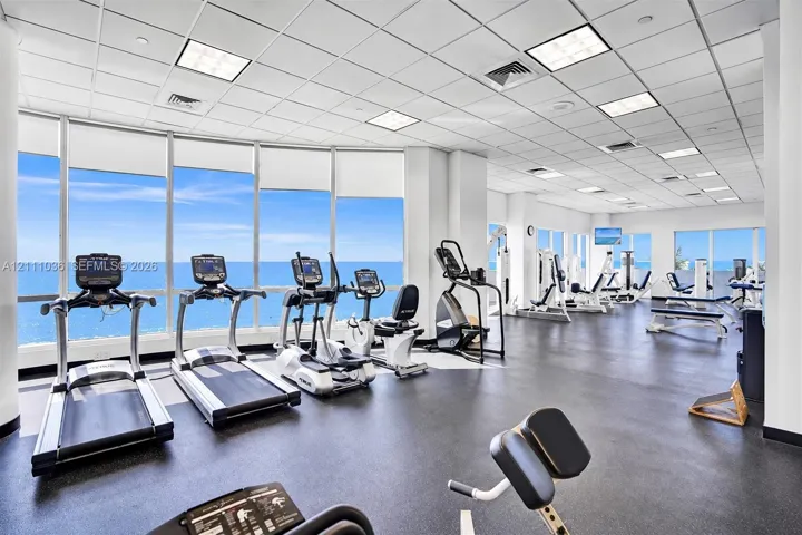 Fitness center featuring expansive floor-to-ceiling windows, panoramic ocean scenery, durable rubber flooring, and a suspended drop-tile ceiling