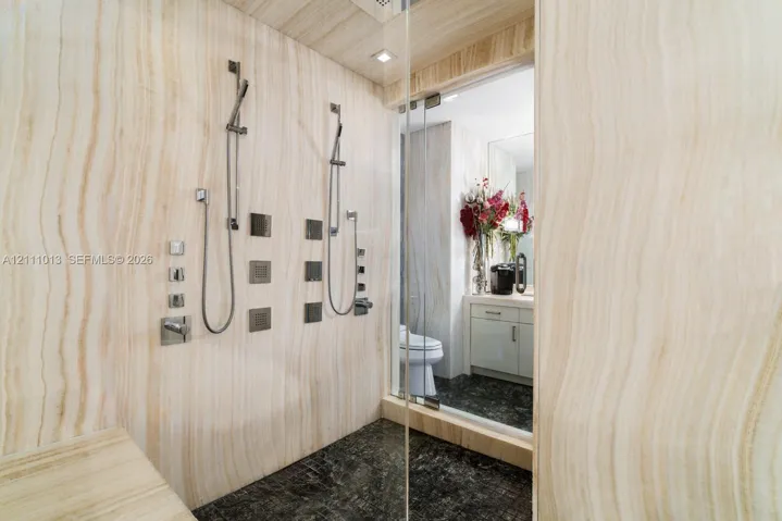 Walk-in shower featuring full-height natural stone wall cladding, dual handheld shower heads with integrated body sprayers, a built-in stone bench, and glass enclosure