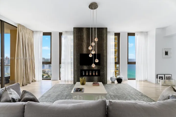 Floor-to-ceiling windows, stone-clad fireplace feature wall, wood-finish flooring, and multi-pendant lighting fixture