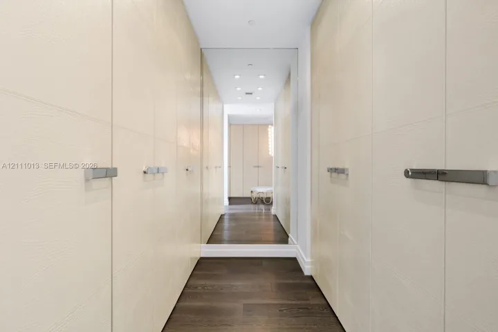 Custom floor-to-ceiling cabinetry with textured cream-colored finish, polished metal hardware, full-height wall mirrors, recessed ceiling lighting, and dark wood-finish flooring