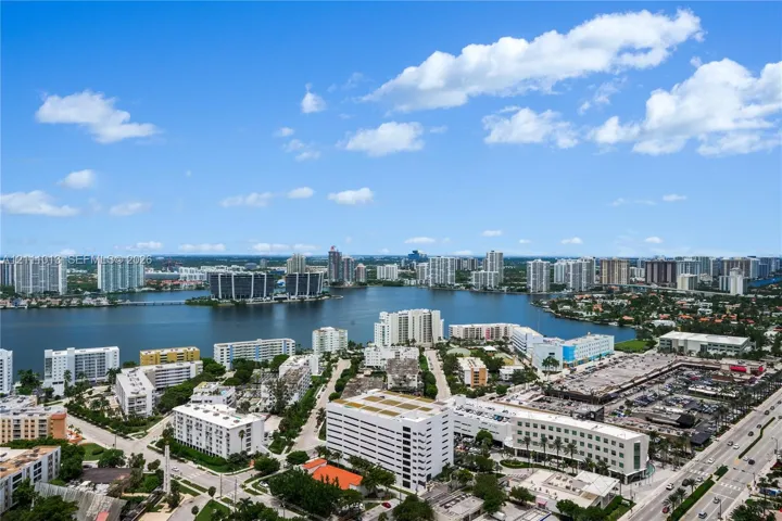 Aerial perspective of a coastal urban landscape featuring multiple high-rise structures, an expansive inland waterway, dense foliage, and established roadway infrastructure