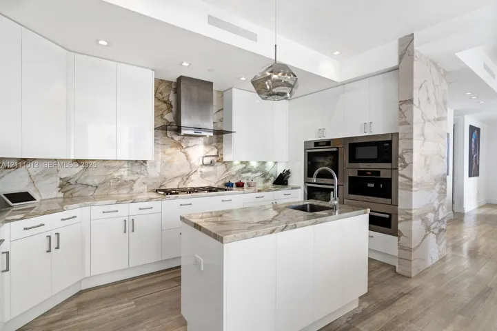 Contemporary kitchen featuring high-gloss white cabinetry, expansive stone slab countertops and backsplash, integrated stainless steel double wall ovens, wood-finish flooring, and a central island with an undermount sink