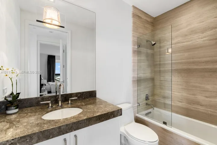 Bathroom featuring a stone-topped vanity with an undermount sink, large wall-mounted mirror, white cabinetry, and a shower-tub combination with a glass panel and stone-finish tile surround