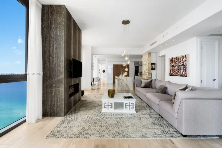 Floor-to-ceiling windows with ocean scenery, prominent dark stone-finished accent wall with integrated fireplace, light-toned wood-finish flooring, island with stone countertop, and multi-pendant light fixture