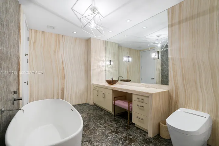 Bathroom featuring floor-to-ceiling stone wall paneling, freestanding soaking tub, custom vanity with integrated stone countertop, vessel sink, and geometric pendant lighting