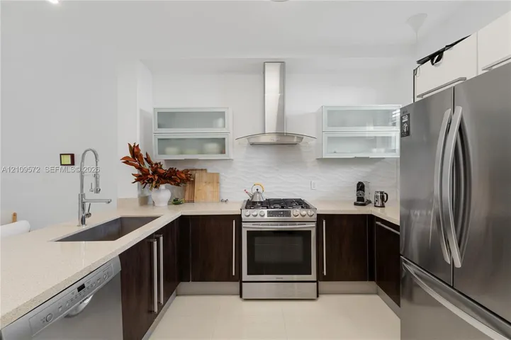 Kitchen featuring dark wood-finish cabinetry, light-toned stone countertops, stainless steel appliances, wall-mounted glass-front cabinetry, and a textured tile backsplash