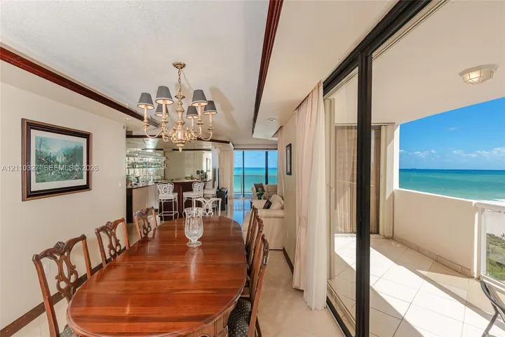 Dining area featuring an ornate multi-arm chandelier, built-in wet bar with mirrored backsplash, and floor-to-ceiling sliding glass doors opening to a tiled balcony with ocean backdrop
