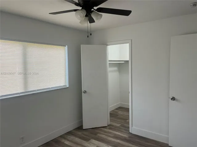 Room featuring wood-finish flooring, a ceiling-mounted light fixture with a fan, a horizontal window with blinds, and a walk-in closet with built-in shelving