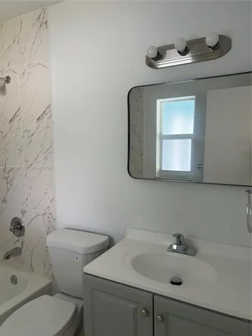 Bathroom featuring a vanity with a white countertop and integrated sink, a rectangular mirror with rounded corners, a three-light vanity fixture, and a bathtub surround with veined tile detailing