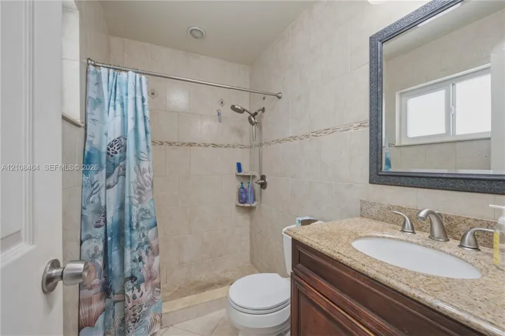 Full-height tiled shower surround with built-in corner shelving, stone-composite vanity countertop, integrated sink with dual-handle faucet, and framed wall-mounted mirror