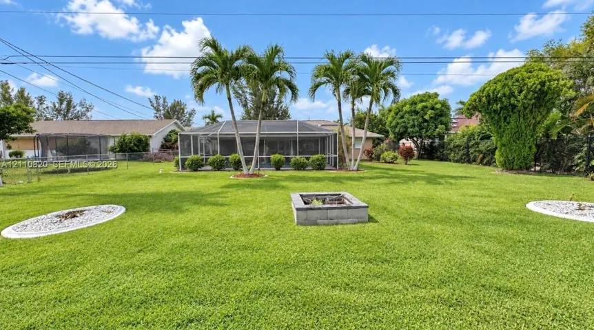 Expansive backyard featuring a screened-in lanai, mature palm trees, a concrete-block fire pit, and stone-accented landscaping beds