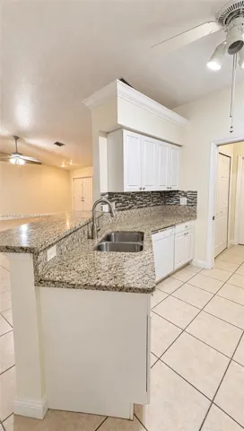 Kitchen featuring a peninsula with a stone-finish countertop, dual-basin stainless steel sink, tile backsplash, white cabinetry with crown molding, and ceramic tile flooring